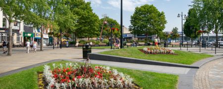 A public square with paved walkways, green grass, flower beds, trees, benches, and shops in the background under a partly cloudy sky.