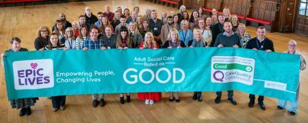 A large group of people stand behind a banner that reads Adult Social Care Rated as GOOD with a Care Quality Commission logo.