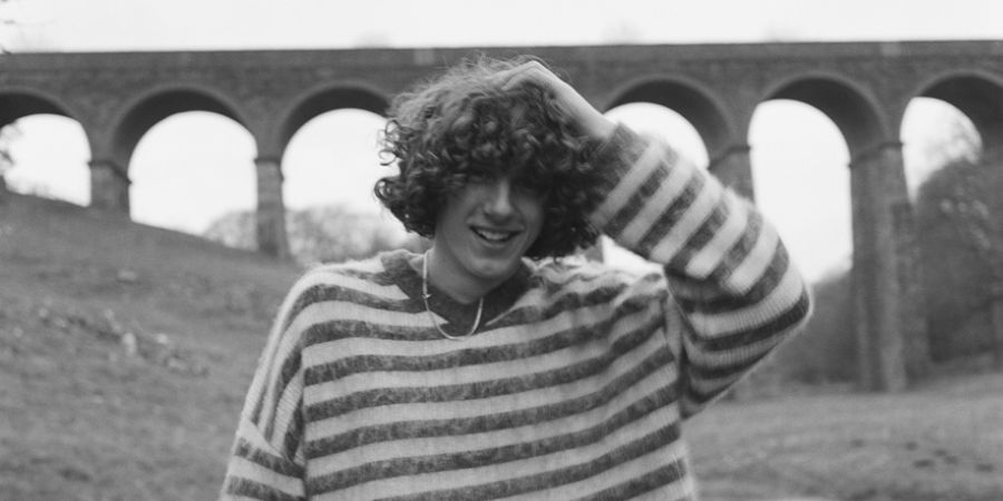 A person with curly hair wearing a striped sweater and a necklace, posing in front of a stone viaduct.