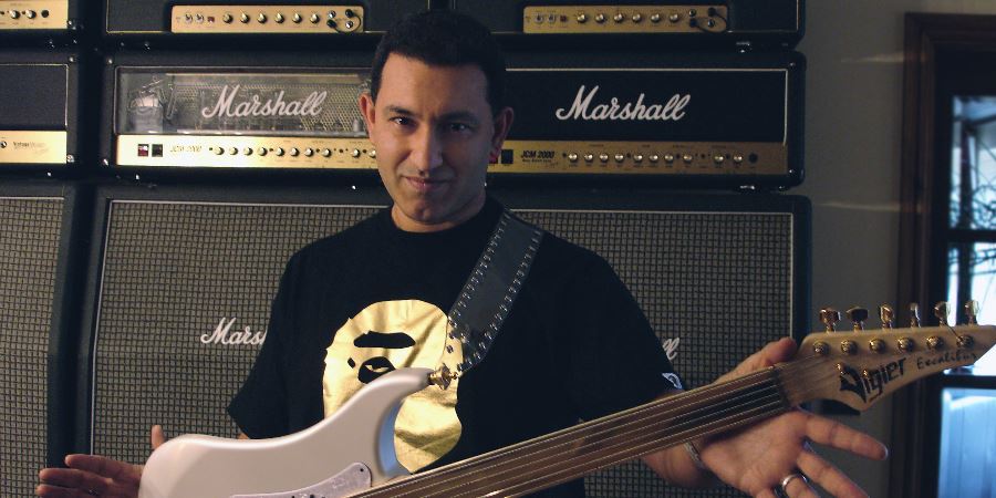 Aziz Ibrahim holding his guitar, stood in front of several amplifiers.