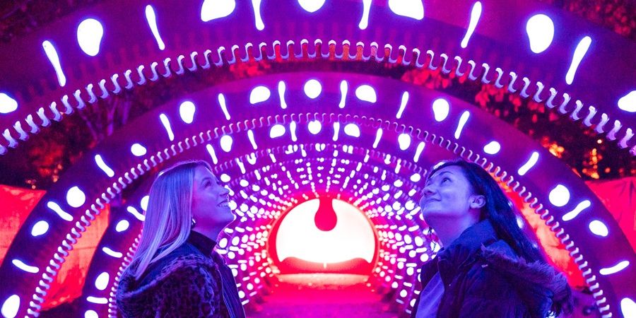 Two people facing each other beneath a bright circular LED tunnel, with glowing purple and white lights forming concentric rings and a central red-and-white spotlight behind them.