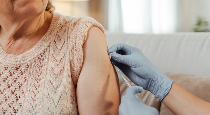 A gloved hand administers a vaccine injection into the upper arm of a person wearing a light pink, open-knit sweater.