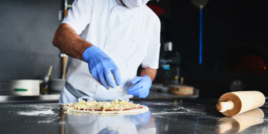 A cook wearing protective gloves and face mask topping a pizza with grated cheese.