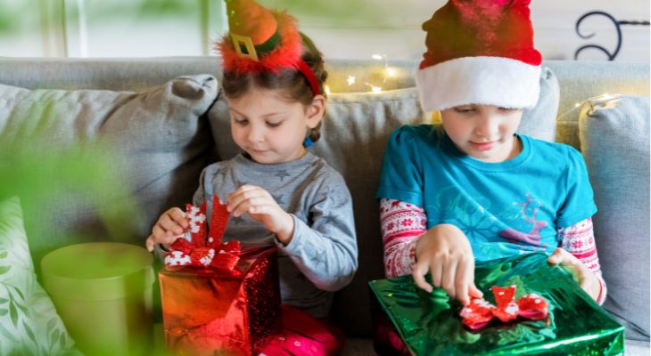 2 children wearing festive hats sat on a grey sofa opening Christmas presents