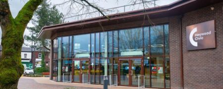 The Heywood Civic building has a modern glass facade with double doors and a brown roof. A sign on the brick wall reads "Heywood Civic".
