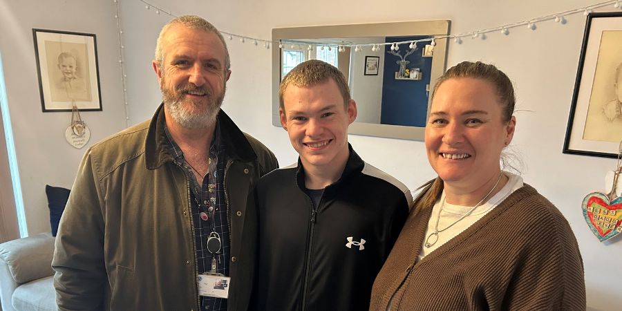 3 people smiling at the camera. A person on the left wears a brown jacket over a patterned shirt. A person in the middle wears a black zip-up jacket with a white stripe. A person on the right wears a brown cardigan over a white shirt.