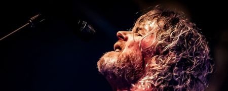 Close-up of a curly-haired performer singing into a microphone on a dark stage.
