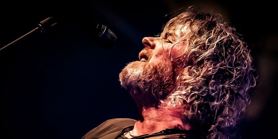 A bearded performer with long curly hair sings into a microphone on a dark stage, lit by warm spotlights.