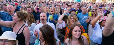 A large crowd of people are at an outdoor event, many with their hands raised in excitement. Some people are holding drinks and wearing festival-style hats.