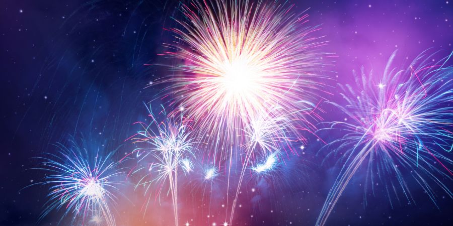Colorful fireworks burst across a dark night sky, with bright white, pink, and blue explosions and sparkling trails fading into a purple-blue haze.