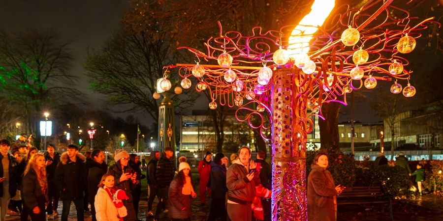 Crowd gathered at night around a brightly lit Christmas tree sculpture with glowing orange and red lights, hanging ornaments, and swirling decorative wires in a park.