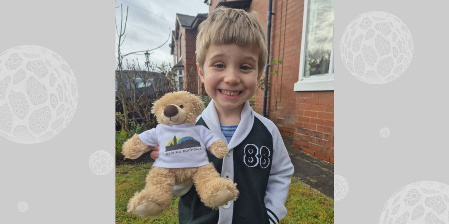 A smiling child holds a teddy bear wearing a white t-shirt with the words "Raising Rochdale" and a silhouette of buildings.
