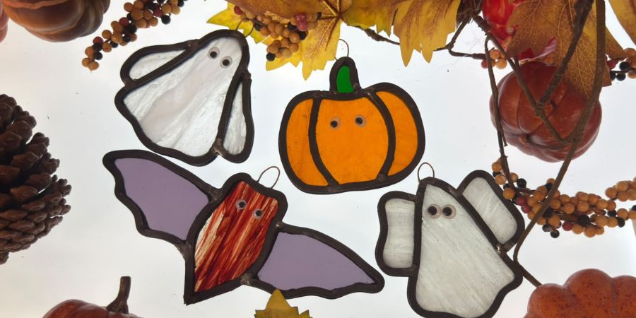 Colorful autumn stained glass figures on a white background: a pumpkin, a ghost, bats, and a small ghost, surrounded by autumn leaves and pinecones with orange and brown tones.