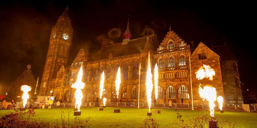 A nighttime shot of the lawn in front of a large gothic building. Large flames can be seen shooting from special pyrotechnic devices.