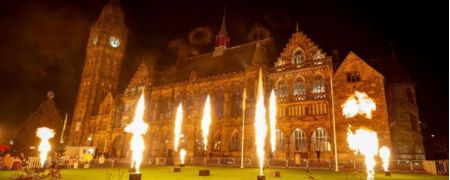 A nighttime view of a large Gothic stone building with tall spires, illuminated by multiple fireworks bursting in front on the lawn.