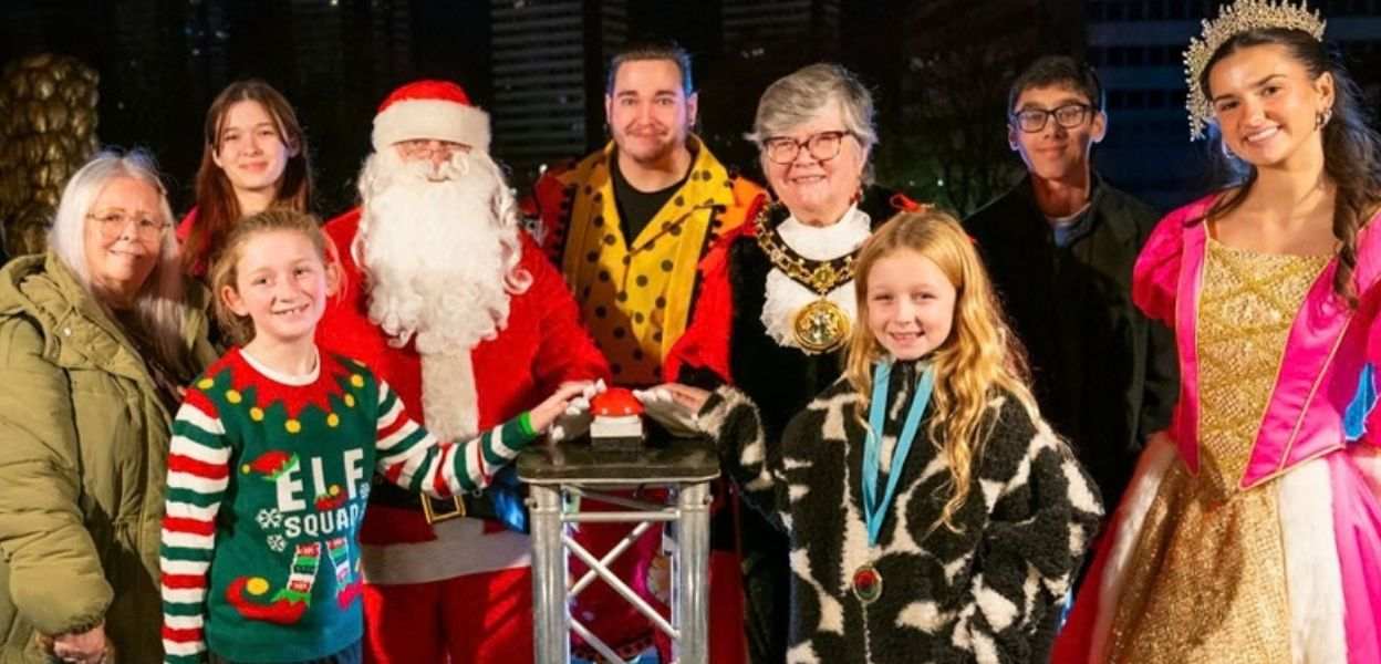 Group photo with Santa in a red suit and white beard beside smiling adults and children in festive outfits,