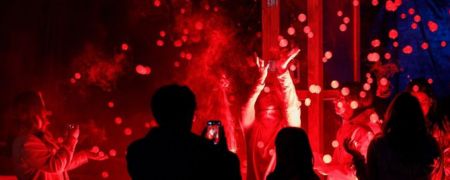 Silhouetted crowd in front of a stage with dancers under intense red lighting, confetti-like particles floating in the air.