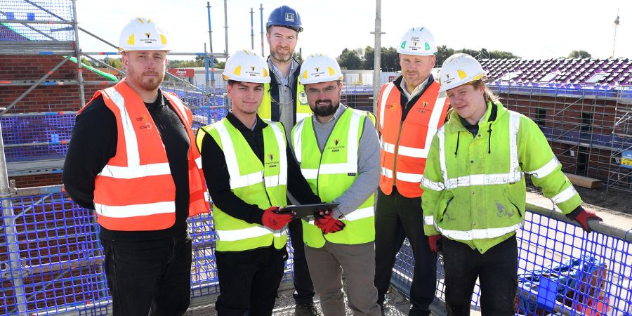 A group of 6 people in high-visibility vests and hard hats stand together on a building site.