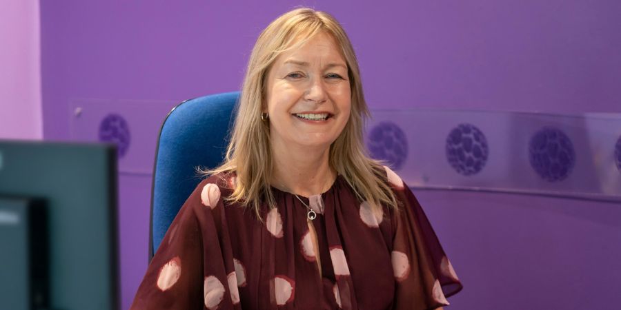 Smiling woman seated in a blue office chair, wearing a maroon top with light pink polka dots, in front of a purple wall with circular patterns and a computer monitor on the left.