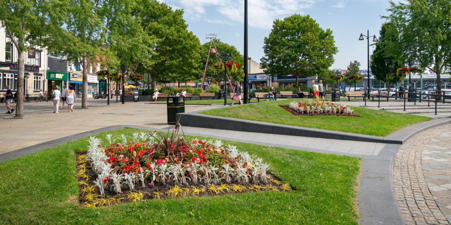 A sunny town square with manicured flower beds, paved walkways, benches, trees, and shops in the background.