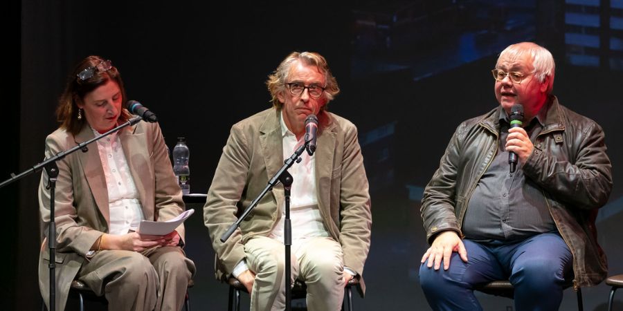 Three speakers sit on stage with microphones: one holding papers at a stand, one seated center with a mic, and one on the right speaking into a handheld microphone under stage lighting.