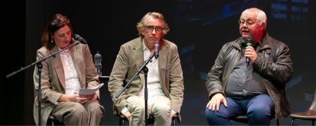 Three speakers sit on stage with microphones: one holding papers at a stand, one seated center with a mic, and one on the right speaking into a handheld microphone under stage lighting.