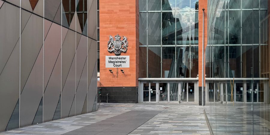 The Manchester Magistrates' Court building features a facade of orange brick and glass, with a crest above the entrance and a modern metal-clad section to the left.