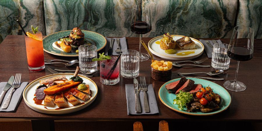 A table setting for a meal with multiple plates of food, drinks, and wine glasses, set against a backdrop of  marbled green seating.