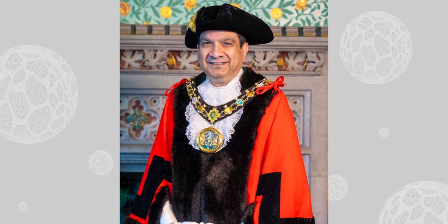 A person in ceremonial robes, a black hat with a plume, and a gold chain of office with a medallion, stands in front of a decorative wall.