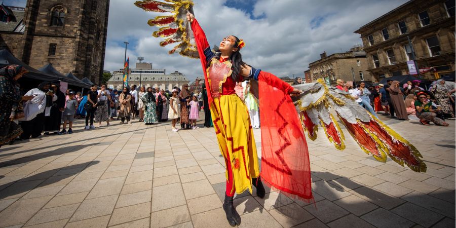 A performer in a colourful costume with large, feathered wings dances in a plaza with a crowd watching and a historic building in the background.