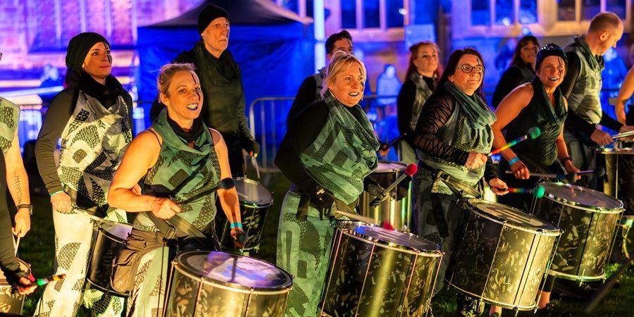 A group of people in matching green-and-white outfits play large silver drums and percussion instruments on an outdoor night stage with blue lighting.