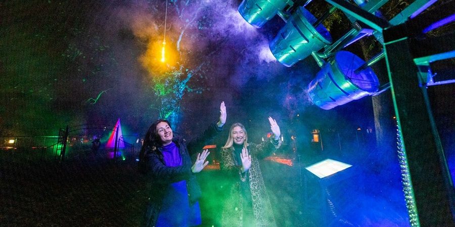 Two performers on an outdoor stage celebrate with raised hands as colorful fog and light beams burst overhead; bright blue and green stage lighting illuminates the scene.