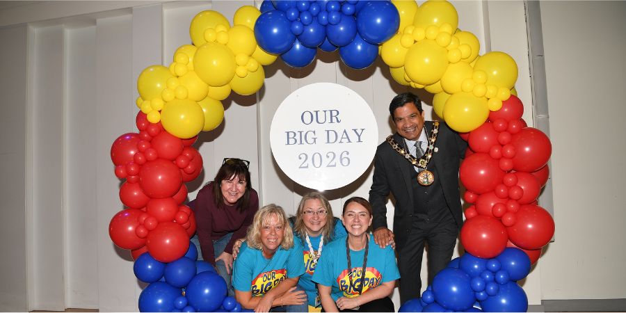 Group of people posing in front of a balloon arch in yellow, blue and red, with a circular sign reading “OUR BIG DAY 2026”
