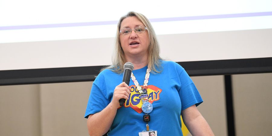 A speaker in a blue T-shirt with a conference badge holds a microphone and addresses an audience in front of a projection screen.