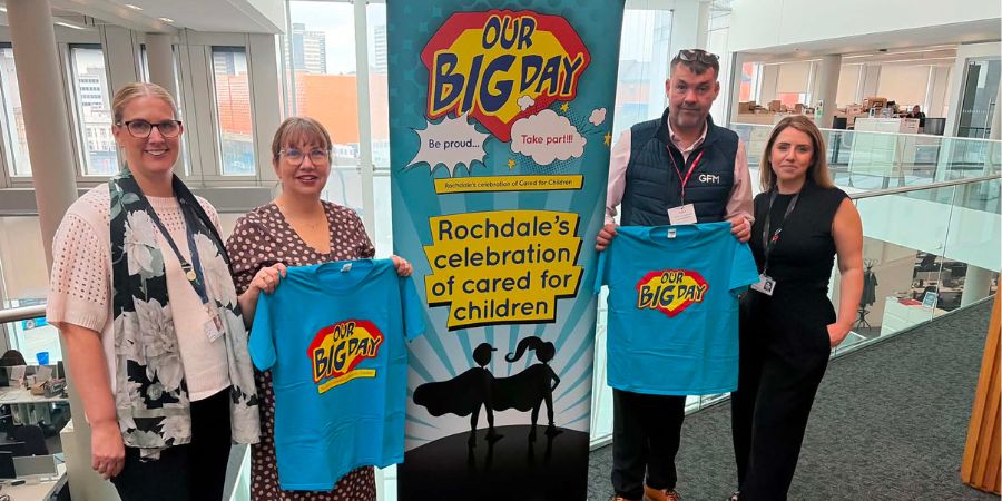 Four people stand in an office holding turquoise “Our Big Day” T-shirts beside a large banner reading “Our Big Day” and “Rochdale’s celebration of care for children.”