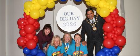 Group posing at a balloon arch in yellow, red, and blue with a circular sign reading “OUR BIG DAY 2026”