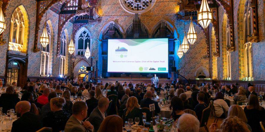 A speaker stands on a stage in front of a large screen in a grand hall filled with people seated at round tables for a formal event.