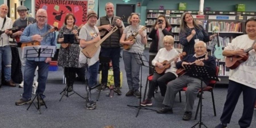 A group of people playing ukuleles in a room with bookshelves in the background.