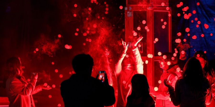 Silhouetted crowd as red stage lighting and swirling confetti-like particles fill the air.