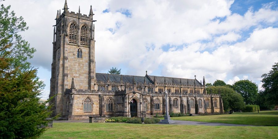 A church with a tall stone tower, arched windows, and a row of buttresses, set on a green lawn under a partly cloudy blue sky.