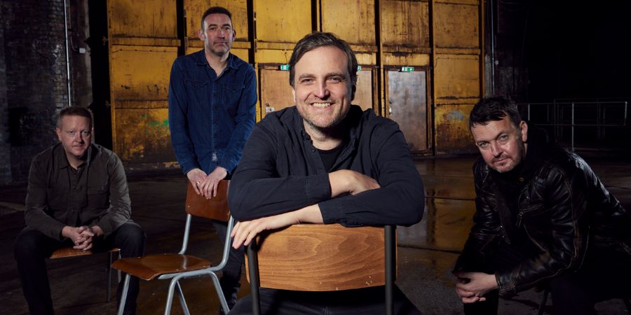 4 men are posing for a photograph in an industrial-looking setting. The man in the centre is smiling and leaning forward on a chair. The other three men are positioned around him.