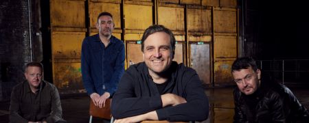 Four men are posing for a photograph in an industrial-looking setting. The man in the center is smiling and leaning forward on a chair. The other three men are positioned around him.
