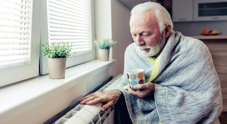 Elderly man wrapped in blanket sat beside a radiator.