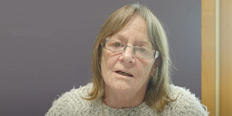 A person wearing a grey jumper and glasses, speaking.