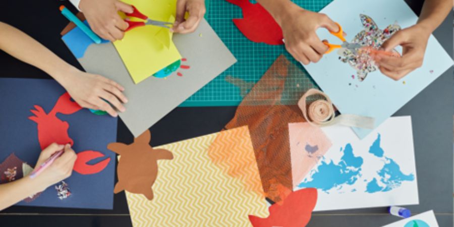 Overhead view of hands cutting and gluing colorful paper crafts, including a red lobster, a brown turtle, and a blue world map.