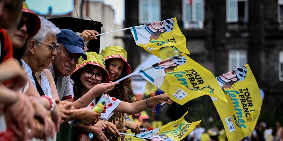 A crowd watches a cycling race, with some people holding yellow flags that say "Tour de France Femmes" and feature a cyclist's portrait. Photo courtesy of A.S.O and Pauline Ballet.