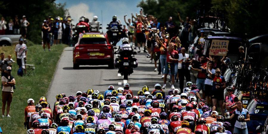 A peloton of cyclists race down a road lined with cheering spectators. A police motorcycle and support vehicles are also present. Photo courtesy of A.S.O and Pauline Ballet.