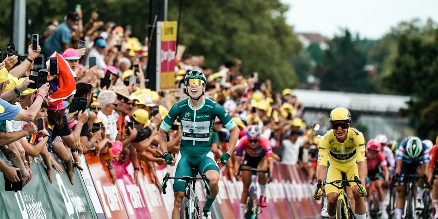 A cyclist in a green racing suit with yellow sunglasses is in the foreground, followed by other cyclists in bright racing attire. Photo courtesy of A.S.O and Thomas Maheux.