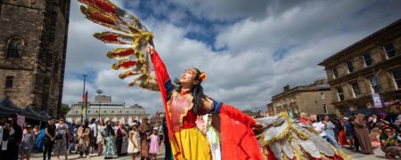 A performer with large, colorful wings dances in a street festival with a crowd watching and historic buildings in the background.
