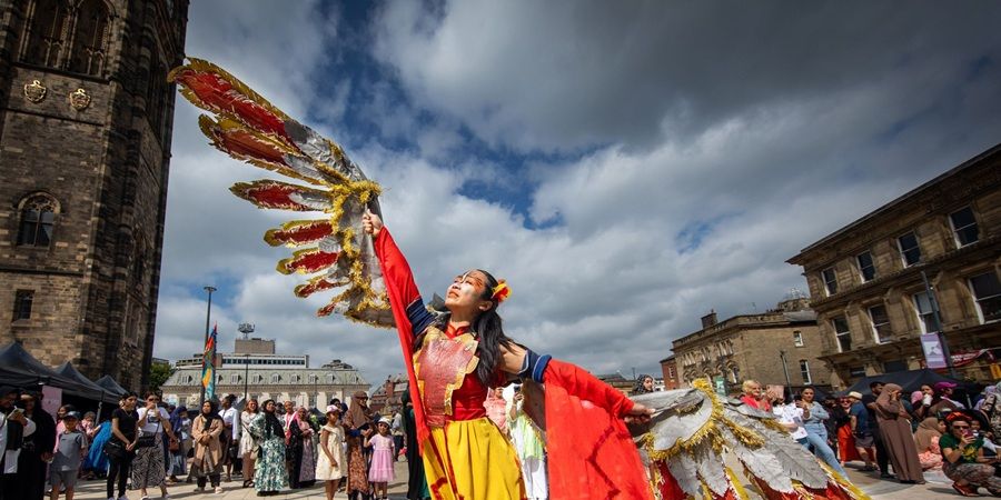 A person in a colorful costume with large, feathered wings performs outdoors in front of a crowd and historic buildings.
